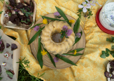 Ein Buffet aus wilden Delikatessen: Ein Gugelhupf dekoriert mit Spitzwegerichblättern und farbigen Wildblumen. Weiter sind Blumen und Blätter in Schokolade und Quark mit verschiedenen Wildkräutern und Wildblumen zu sehen.