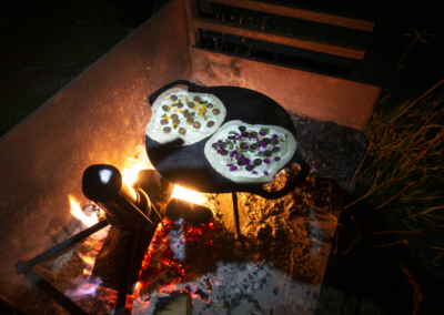 Ein Feuer lodert, süsse Flammkuchen mit Schokolade und Wildblumen backen auf einer Grillplatte, daneben steht eine italienische Kaffeekanne.