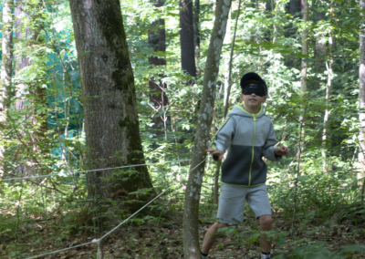 Orientieren: Ein Kind folgt mit verbundenen Augen einem Seil durch den dichten Wald, an welchem ca. einen Meter vor dem Kind ein Stoffbeutel hängt.